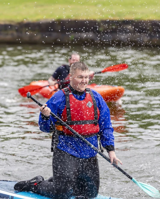 Young man kneeling on a paddleboard, holding a paddle and smiling as water splashes around him. Young man kneeling on a paddleboard, holding a paddle and smiling as water splashes around him.